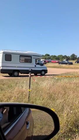 The video shows a white camper van driving on a dirt road on a sunny day. The camper van is moving from left to right across the frame. In the foreground, a portion of a car's side mirror is visible, suggesting the video is being filmed from inside another vehicle. The environment appears to be a grassy field with some trees and other vehicles in the background. There are some yellow and red tapes along a wooden post in the foreground, indicating a possible boundary or demarcation. The sky is clear and blue.