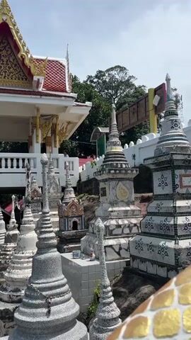 The video pans across a collection of small stupas, which are funerary monuments. Some are white and conical, while others are more elaborate, adorned with colorful tiles and decorative patterns. These stupas are arranged in rows on a rocky, overgrown hillside. Behind them, a building with a red-tiled roof and gold trim is visible. Lush green trees and a bright blue sky with scattered clouds form the backdrop. A vibrant red flowering tree adds a splash of color to the scene. The overall impression is of a tranquil, perhaps spiritual, outdoor space.