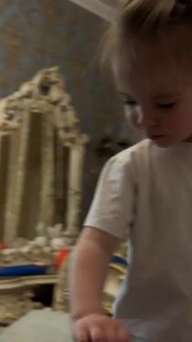 A young child with light brown hair, styled with a green clip and a dark blue barrette, is captured in a series of close-up shots. The child is wearing a simple white t-shirt and appears to be in a bedroom. The background features ornate, gold-accented wallpaper and a large, antique-style mirror with carved detailing. A chandelier or decorative light fixture is visible on the wall. In some frames, the child is looking down, while in others, they are looking directly at the camera with a slight smile. The child's body language suggests playful movement, with arms extended in one shot. The overall impression is one of a candid moment of a child at play in a richly decorated room. No visible text is present in the media. The location context is Ufa, Russia.