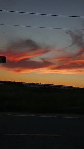 A video capture of a sunset viewed from a roadside. The sky is a blend of deep blue, orange, and fiery red, with dramatic clouds painted in shades of pink and orange. A dark silhouette of a field and a distant line of buildings stretches across the horizon. In the foreground, a paved road is visible, with a thin white line marking the lane. Overhead, power lines are faintly discernible against the darkening sky. The scene is static, with no discernible movement.