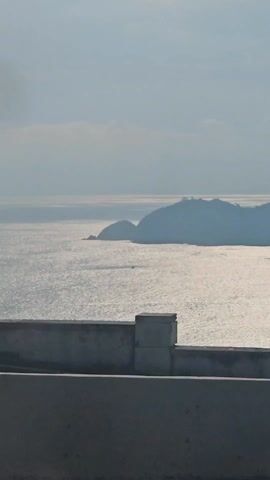 The video shows a view of a body of water with a distant island. The water is shimmering in the sunlight. In the foreground, there is a low concrete barrier. As the video progresses, the camera pans to reveal several flags waving in the wind. The flags appear to be various national flags. The camera continues to pan, showing palm trees and parking signs in the foreground, with the water and island still visible in the background. The video seems to be taken from a moving vehicle.