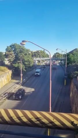 The video shows a view from above looking down at a road with traffic. The road curves slightly to the right and goes under an overpass. There are trees on the left and right sides of the road. The sky is clear and blue. There are several vehicles on the road, including cars, a white van, and a motorcycle. There are also streetlights lining the road. In the foreground, there is a yellow and black striped barrier.