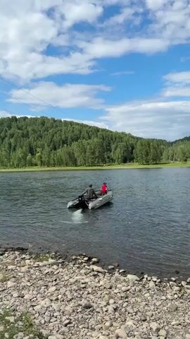 A small inflatable boat with two people is moving across a wide, calm river. The person in the back appears to be steering the boat with an outboard motor, which is creating a white wake behind the vessel. The person in the front is facing forward. The riverbanks are lined with smooth, grey stones. In the background, a lush, green forest covers rolling hills. The sky is blue with scattered white clouds, and a power line is visible stretching across the upper left corner of the frame. The overall impression is a peaceful, natural outdoor scene.
