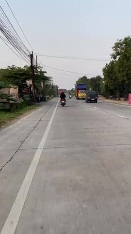The video shows a POV shot from a motorcycle traveling on a paved road. The road has a white line dividing the lanes. The rider is wearing a red helmet and a dark jacket. There are other motorcycles and vehicles on the road. The environment is rural, with trees and vegetation on the sides of the road. There are also power lines and poles. The sky is overcast. The video is in color and appears to be filmed during the daytime.