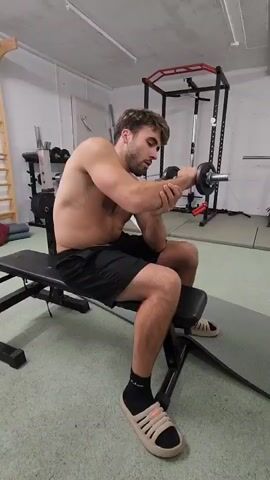 Here is a description of the video:
A shirtless man is sitting on a bench in a home gym, with his right forearm resting on his left thigh. He is holding a dumbbell in his right hand and performing wrist curls. The man is wearing black shorts, black socks, and beige sandals. A weight rack, a barbell, and a workout machine are behind him.