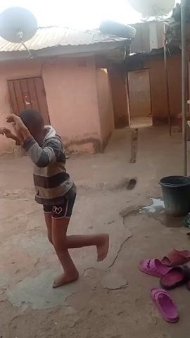 A young boy is dancing barefoot in a courtyard. He is wearing a striped long-sleeved shirt and shorts with a heart on them. He moves his arms and feet to an unheard rhythm, focusing intently on his movements. The courtyard is paved with cracked concrete. In the background, there is a pinkish-brown building with a tin roof, and a doorway leading to another area. A satellite dish is mounted on the roof. A black bucket sits to the right of the frame. A pair of pink sandals and a pair of dark sandals are on the ground near the bottom of the frame.