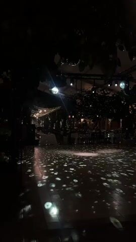 The video displays a dark outdoor scene at night, possibly a reception or event area. Disco balls are suspended from above, casting circular patterns of light onto the tiled floor. Tables and chairs are visible in the background, suggesting a dining or gathering space. String lights are draped across the area, adding to the festive ambiance. The foreground is dominated by the reflective patterns of light on the floor, with silhouettes of trees and foliage framing the scene.
