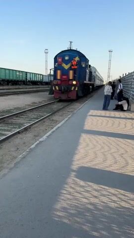 A blue and yellow train is stopped at a station platform. A person in an orange vest is standing on the front of the train, looking out. Several people are standing on the platform, some appear to be waiting for the train. The platform is paved and there are train tracks running alongside it. The sky is clear and blue. The train appears to be a passenger train. The people on the platform are dressed casually. The person on the train is wearing work clothes. The train has several carriages behind the locomotive. The overall impression is of a typical train station scene.
