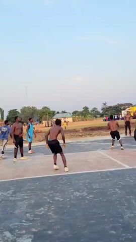 The video shows a group of young men playing basketball on an outdoor court. The basketball court is marked with lines and is located in an area with a dirt field adjacent to it. In the background, there is a building, trees, and other people. The players are dressed in shorts, some without shirts, and athletic attire. They are actively engaged in a basketball game, with one player shooting at the hoop, and others trying to rebound the ball. There is a towel hanging from the basketball hoop structure. The sky is blue and the lighting suggests it is daytime.