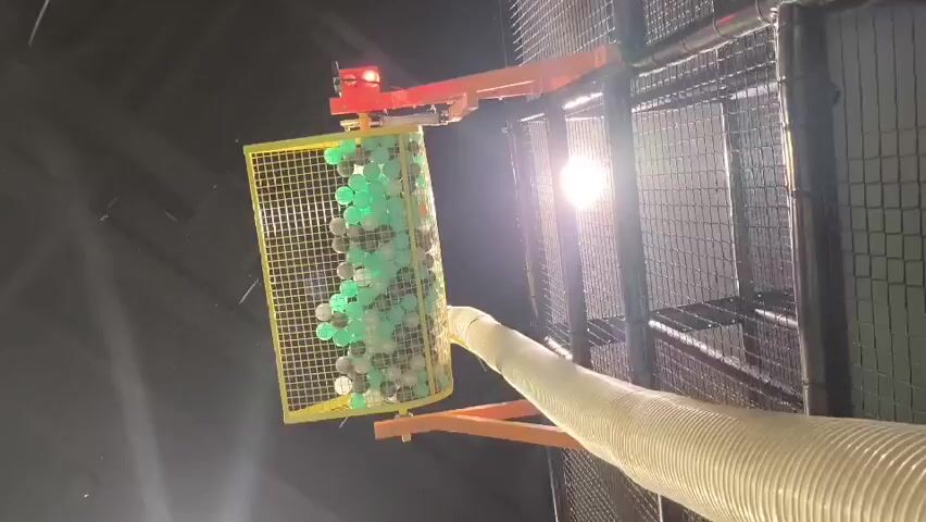 This video shows a ball pit filling up with balls from a tube. The balls are various colors, including green, white, and grey. The ball pit is contained in a yellow cage with a metal frame. The cage is elevated, and a light source is visible in the background. The balls are being ejected from a large, flexible tube into the cage. The video appears to be taken from a low angle, looking up at the ball pit.