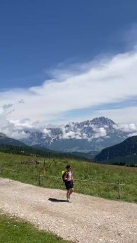 A vertical, handheld video pans across a scenic mountain landscape. The first frame shows a person with a yellow backpack walking on a dirt path, with a green grassy field and majestic mountains in the background. The mountains are partially covered in clouds. The path has a low fence made of posts and wire. The video then pans to the left, showing more of the path curving away and the mountain range continuing, with some trees visible on the lower slopes. The next frame shifts focus to a prominent, jagged rock formation towering over a building nestled on a green hillside. People can be seen walking on the path in the distance. The final frames zoom in on the building, revealing it to be a rustic lodge or restaurant with stone and wood construction, situated in front of the same striking rock peak. There are outdoor seating areas with tables and umbrellas, and people are visible sitting and walking around the building. The sky is a clear, bright blue throughout the video.