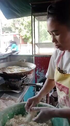 A woman wearing a white t-shirt and a yellow apron is in a food stall. She is standing in front of a large wok filled with boiling oil, where pieces of food are being fried. Her hands are covered in a white powdery substance, and she is mixing a ball of dough in a large green bowl. The stall appears to be outdoors, with a glimpse of trees and buildings in the background.