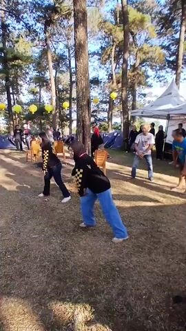 A group of people are outdoors participating in some kind of group activity or exercise. The setting appears to be a campground or park with tall trees and grassy ground. Several tents and white canopies are visible in the background, suggesting an organized event or gathering. In the foreground, three women are positioned in a line, facing towards the right of the frame. They are all wearing similar black tops with a yellow checkered pattern on the sleeves, and blue pants. Their body language suggests they are engaged in a coordinated movement, possibly a dance or stretching routine, with their arms extended to their sides and bodies leaning forward. Further back, several other individuals are also visible, some appearing to be following along with the activity. The lighting suggests it is daytime, with sunlight filtering through the trees. Yellow lanterns are hanging from the trees, adding a festive touch to the scene.