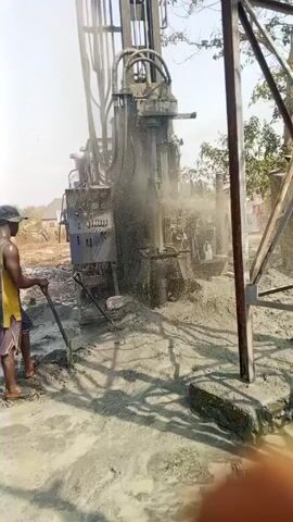The video shows a large drilling rig at a construction site, with one man standing nearby with a shovel. The rig is churning up mud and spraying water. The man stands close by. The ground around the rig is covered in mud. The sky is bright. Trees are visible in the background.