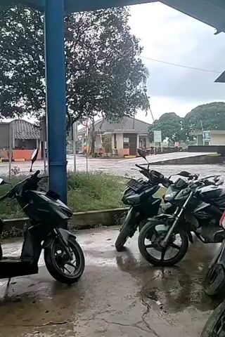 The video captures a scene with several motorcycles parked outdoors. In the background, a building with a newly tiled roof is visible, suggesting repair or construction work. Several people are seen near the building, some appearing to be workers on ladders or observing the progress. Stacks of roof tiles are piled near the building, indicating the materials used for the roofing project.

The setting seems to be an open area, possibly a parking lot or a yard associated with the building. The weather appears overcast, with a cloudy sky, and the ground looks wet, suggesting recent rain or damp conditions. The time of day is not explicitly clear, but the lighting suggests it's daytime.

Notable details include the presence of multiple motorcycles, a prominent building undergoing roofing work, and several individuals involved in or observing the construction. The location cues point to Cotgirek, Indonesia. There is no clear indication of emotions being conveyed, and the overall mood appears neutral and focused on the ongoing activity.

Visible text is limited to license plates on some of the motorcycles. One plate clearly reads "BL 3935 KAV" and another "BL 3999 PAS". There is also some text on a sign above the building, but it is too blurry to read. The presence of air conditioning units on the building might suggest it's an office or a commercial establishment. The scene gives the impression of a functional day with ongoing maintenance or construction work.