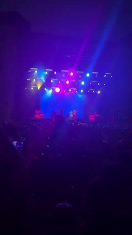 The video shows a live music concert from the perspective of the audience. The stage is illuminated with bright purple and yellow lights, with a large blue screen serving as the backdrop. A band is performing on stage, with three visible members. One guitarist stands on the left, silhouetted against the lights. In the center, a lead singer is on his knees, gesturing with his hands and looking up. Another band member stands behind him, holding a guitar. The audience in the foreground is largely silhouetted, with some hands raised and one person holding up a phone to record. The atmosphere is energetic and festive, with the vibrant lighting and the crowd's engagement indicating an active performance.