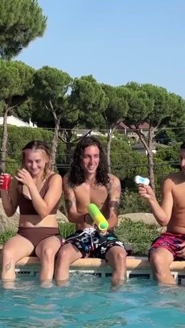 Three young adults are sitting on the edge of a swimming pool. The person on the left, a woman with blonde hair, is holding a red cup and laughing. The person in the middle, a man with long, dark curly hair, is holding a green and yellow water gun and looking at the woman. The person on the right, a man, is holding a small white object and smiling. Water splashes into the pool from the left. The background shows trees and houses.