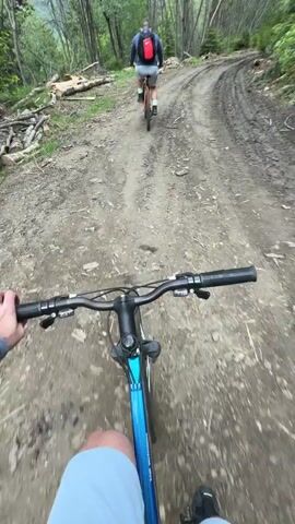 The camera perspective shows two people mountain biking on a muddy, dirt road in a forest. The camera is attached to the handlebars of the bike in the rear. The second biker rides ahead of the camera perspective and has a red backpack. Fallen logs and tree branches are visible along the sides of the road. The trees on either side of the road are tall and mostly bare of leaves. The rider of the camera bike is wearing light blue shorts.