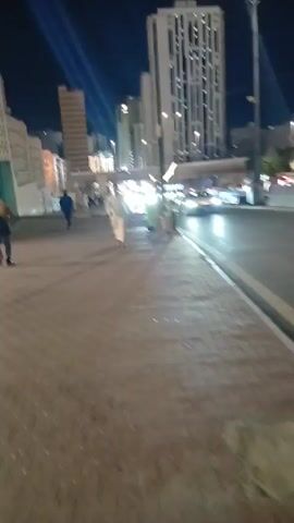 Here is a description of the video:

The video shows an urban scene at night. A street with car traffic runs alongside a sidewalk paved with bricks. Pedestrians are visible on the sidewalk and in the distance.  Buildings of varying heights are illuminated and line the street. Streetlights illuminate the area, and in the background, beams of light project into the night sky.  A white car drives by on the street, followed by other vehicles.