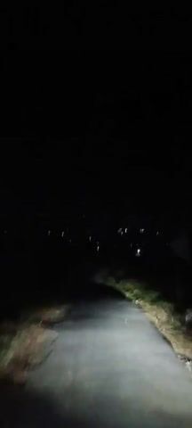 The video shows a first-person perspective of someone driving down a narrow, paved road at night. The road is bordered by grass and vegetation on either side. In the distance, there are scattered lights, suggesting a town or settlement. The headlights of the vehicle illuminate the road ahead, casting shadows and highlighting the rough texture of the pavement. The sky is dark, indicating it is nighttime, and there are no visible stars or moon. The overall atmosphere is one of isolation and darkness.