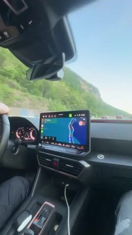 The video is shot from inside a car, likely from the front passenger seat, showing the car's interior and the view through the windshield while driving on a highway.

The interior of the car features a modern dashboard with a prominent, large central infotainment screen. The screen displays a navigation map, with various icons and text, including a visible time of "18:56" and a temperature of "23.0°". Red trim or stitching accents the top of the dashboard. A phone charging cable is seen connected in the center console area. Parts of the driver's and passenger's legs, dressed in jeans, are visible in the lower frame. The rearview mirror and the driver's side mirror are also visible, reflecting the sky and surrounding landscape.

Outside the car, a multi-lane highway stretches ahead. The landscape is mountainous, with green, tree-covered slopes rising in the distance on both sides of the road. Protective barriers line the side of the highway. Other vehicles are visible on the road ahead. The sky is clear and light blue, with a single, faint contrail visible. The lighting suggests it is late afternoon or early evening. The scene depicts travel through a scenic, mountainous region, consistent with Val Mara, Switzerland.