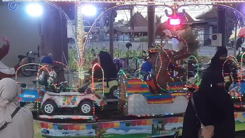 A carousel ride for children is in motion. The ride features small, brightly colored vehicles, resembling jeeps and cars, adorned with numerous LED lights. A baby wearing a blue hat is seated in one of the vehicles, appearing to be the sole occupant. Several people are visible in the foreground, mostly women wearing hijabs, some observing the ride, and one woman is taking a photograph with her phone. The background shows a street scene with buildings and vehicles, suggesting an outdoor public space, possibly a market or fairground, during daylight hours. The carousel itself is illuminated with a variety of colored lights, creating a festive atmosphere.
