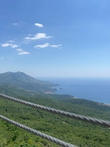 The video shows a panoramic view of a coastal landscape from a high vantage point, likely a mountain. The foreground is dominated by two thick, metallic cables running horizontally across the frame, suggesting a cable car or a viewing platform with safety railings. Beyond the cables, a lush green, forested hillside slopes downwards towards a body of water, which appears to be the sea. Along the coastline, a town or city is visible, with buildings clustered near the shore and extending inland. The water is a deep blue, meeting a clear, bright blue sky with a few wispy white clouds. The overall impression is one of a scenic and expansive vista on a clear, sunny day.