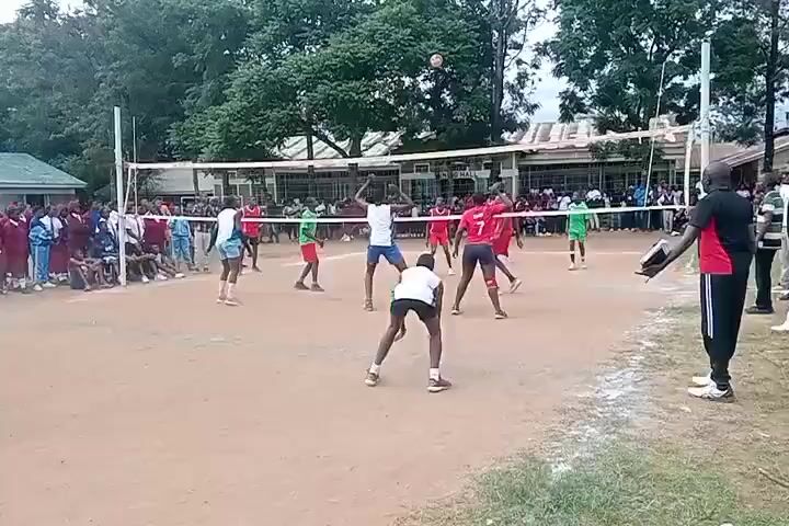 The video shows a volleyball game taking place outdoors on a dirt court. Two teams of young women in athletic wear, one in red uniforms and the other in teal and white, are actively playing. A net divides the court, and spectators, mostly young people in school uniforms, are seated and standing along the sidelines. A coach or official dressed in black pants and a red and black top stands on the side, observing the game. The background includes trees and buildings. The camera angle is static and provides a clear view of the entire court.