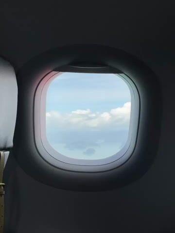 A view through an airplane window shows a tropical seascape with islands and clouds. The window is a rounded square shape and is framed by the dark interior of the aircraft. The sea is a calm blue-green, and several small islands with lush vegetation are visible. Wispy white clouds dot the bright blue sky. The camera is stationary, offering a static perspective of the landscape.