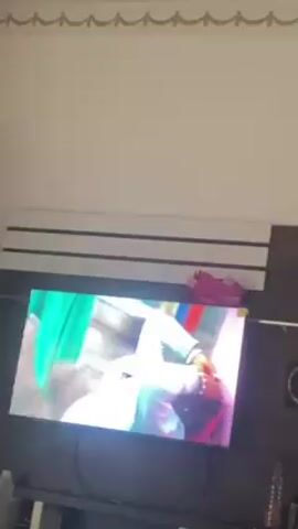 A television set displays a music video in a room. The video features a Black man with a pink hat lying in a hammock, and a Black woman in a white top and patterned skirt dancing. The room has white walls with gold trim near the ceiling and a white and black striped shelf above the television. There are pink shoes and various objects on the shelf and the TV stand.
