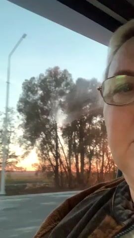 The video shows a person with blonde hair and glasses, partially visible in the lower right corner. They are wearing a brown jacket. The person appears to be inside a vehicle, looking out of a window. The background shows a sunset over a flat landscape with trees and a streetlamp. The sky is a mix of blue and orange hues.