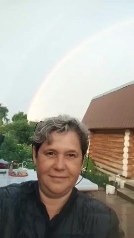 A woman with short, grey hair and a black jacket is taking a selfie outdoors. Behind her, a rainbow arcs across a pale sky. To her right is a wooden house with a brown roof. The ground appears wet, suggesting it has recently rained. In the background, there are trees and other buildings.