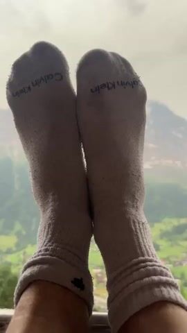This media file captures a serene moment with a person's feet, clad in Calvin Klein socks, resting in the foreground. The socks appear to be a light gray color and show some signs of wear. Behind the feet, a breathtaking panoramic view of the Swiss Alps unfolds. The scene is set in Grindelwald, Switzerland, characterized by its iconic mountainous landscape. 

The backdrop features majestic, snow-capped mountains under a cloudy sky, suggesting a possibly cool or overcast day. Below the towering peaks, a lush green valley is dotted with a charming village, complete with houses and verdant fields. The overall impression is one of peaceful contemplation and appreciation for the natural beauty of the Swiss Alps. There are no specific activities or events clearly depicted, other than the relaxed posture of the person enjoying the view. The visible text on the socks reads "Calvin Klein".
