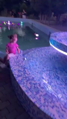 A young girl with blonde hair in a bun, wearing a pink t-shirt and patterned shorts, sits on the tiled edge of a fountain. The fountain has blue and white mosaic tiles and is illuminated with purple and blue lights. The water in the fountain has colorful lights reflecting on its surface, with a few small water jets visible. The girl is looking towards the fountain, and her arms are outstretched, as if interacting with something. She appears to be happy and engaged. In the background, there are trees, and a few other people are visible in the distance, walking around. String lights are also visible in the trees, suggesting it might be evening. The overall setting is an outdoor park or recreational area.