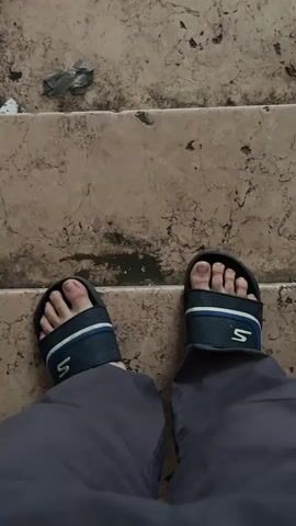 The video begins with a close-up shot of a person's feet wearing dark grey pants and blue and white slide sandals. The camera is positioned looking down at the feet, which are standing on a set of stone steps. The steps are worn and appear dirty with some debris scattered on them. The person's feet are bare inside the sandals. The camera then moves upwards slightly, showing more of the steps and the debris. The scene then abruptly cuts to a view of a broken window frame. Through the window, an outdoor courtyard or alleyway is visible. The walls of the courtyard appear to be made of a textured, light-colored material. There is some debris on the ground visible through the broken window.