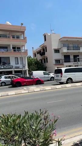 This video captures a street scene in Paphos Municipality, Cyprus, on a bright, sunny day. The time of day appears to be mid-morning to early afternoon, indicated by the high sun and clear blue sky.

The scene is a busy street with several vehicles moving. Prominently featured is a sleek, bright red sports car, possibly a Lamborghini, driving on the right side of the road. In the foreground, a white SUV partially obscures the view. Other vehicles visible include a white van, a dark-colored minivan, a maroon sedan, and other cars.

The buildings lining the street are typical of a Mediterranean coastal town. They are multi-story structures, likely apartments or shops, with balconies and light-colored facades. One building on the left has signage that reads "sh shalore store". Another building on the right has signs that appear to be for a bakery, with Greek text that includes "ΦΟΥΡΝΟΣ".

The activity depicted is simply the flow of traffic on a street. There are no distinct events or organized activities taking place. The overall mood is one of a typical day in a sunny, developed urban area. The vegetation in the foreground suggests a well-maintained public space with landscaping. The presence of multiple cars and commercial establishments points to a commercial or residential district.