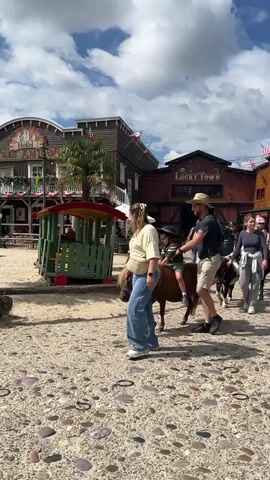 The video shows a sunny day in what appears to be an amusement park or themed village with Wild West-style buildings. A group of people are walking on a gravel path. In the foreground, a woman in a yellow shirt and blue jeans walks to the left, leading a small brown pony with a child on its back. Behind her, a man in a straw hat, dark t-shirt, and khaki shorts walks to the right, holding the pony's reins. The child on the pony is wearing a dark hat. Further back, other people are visible, including a woman in a grey top and white pants walking a black pony with a child on it, and another person leading a black pony. In the background, wooden buildings with signage such as "Hotel Red Auroro" and "Lucky Town" are visible, along with palm trees and a bright blue sky with white clouds. The ground is covered in gravel and some scattered stones. The overall atmosphere is casual and leisurely.