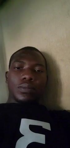 The video is a close-up shot of a Black man. He's wearing a black t-shirt with a white design. He is leaning against a beige wall with textured paint. The lighting is somewhat dim. He is looking directly at the camera.