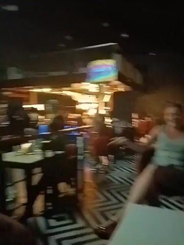 The video shows the interior of a dimly lit restaurant or bar. There are people seated at tables and others walking around, including service staff. The furnishings include dark chairs and tables, and there is a large chandelier hanging from the ceiling. A screen in the background displays colorful geometric patterns. The overall atmosphere appears lively and social.