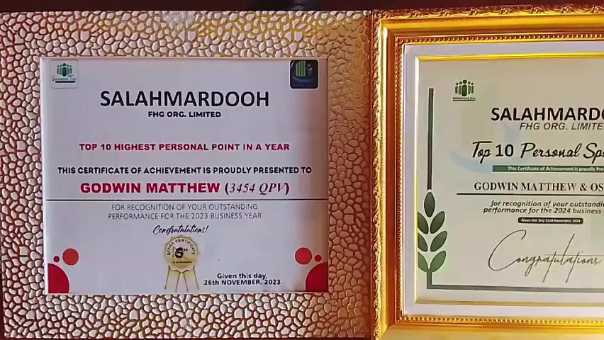 The video shows two framed certificates from the company "SALAHMARDOOH FHG ORG. LIMITED." Both certificates are for "Top 10 Personal Sponsoring." The certificate on the right is awarded to "GODWIN MATTHEW" for his outstanding performance for the 2023 business year. It indicates he placed 3rd. The certificate on the left is awarded to "GODWIN MATTHEW & OSHI (9)" for their outstanding performance for the 2024 business year, and indicates they placed 1st. The certificate on the right is dated "26th NOVEMBER, 2023." Both certificates have a gold frame. The frame on the right has a smoother finish, while the frame on the left has a pebbled or textured surface.