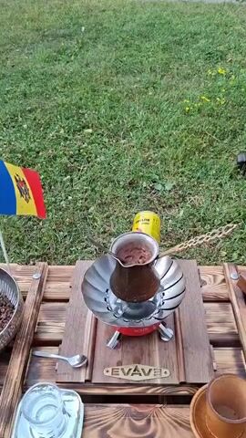 This is a vertical video recorded outdoors in a grassy area with trees in the background. In the foreground, a small portable camping stove is set up on a wooden table. On top of the stove is a cezve (a small pot used for brewing coffee) filled with what appears to be coffee grounds and liquid. Next to the cezve, a can labeled "BUTANE" is visible. A Moldova flag is planted in the ground to the left of the stove. Further back, on the grass, are several military aircraft, including helicopters and airplanes, suggesting the location might be an open-air museum or a historical site. One of the planes has a large, tall missile-like object mounted vertically next to it. The sky appears overcast, indicating it could be late afternoon or early evening. The overall atmosphere seems relaxed, with the activity of preparing coffee in a natural setting, juxtaposed with the imposing military hardware. The visible text is "BUTANE" on the gas can and "EVAVE" on the wooden table support. The timestamp "00:00" is also visible at the beginning of the video, indicating the start of the recording.