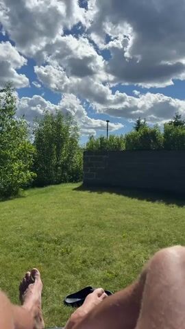 The video shows a first-person view of a person relaxing outdoors on a sunny day with fluffy clouds in the sky. In the foreground, the person's bare legs and feet are visible, with a pair of black flip-flops lying on the grass next to their right foot. The scene is a lush green lawn, bordered by trees and a low brick wall in the background. A tall pole with a light fixture is visible behind the wall. The camera appears to be stationary, offering a peaceful, unmoving perspective of the outdoor environment.