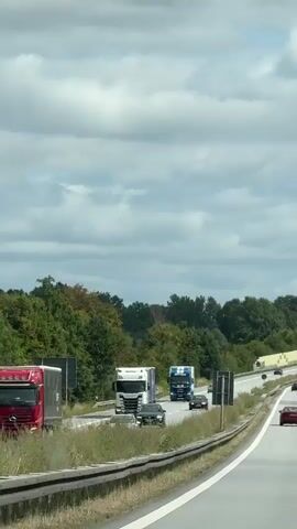 This is a vertical, first-person video taken from a vehicle moving on a highway. The road is paved with white lines dividing the lanes. There are several cars and two trucks on the highway. The truck on the left is white and blue, and the truck on the right is blue and white. The cars are mostly red and black. The highway is lined with trees on the right side, and there is a sound barrier with a yellow and white patterned design. The sky is cloudy. The timestamp indicates the video lasts for approximately 4 seconds.