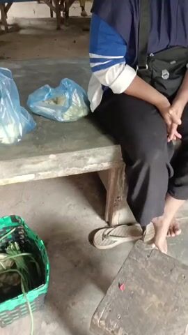 A woman wearing a hijab and a blue and white striped shirt sits on a bench in what appears to be an outdoor market. She is wearing black pants and is barefoot. To her left is a blue plastic bag. To her right, on the bench, is another bag. She is looking to her right. The background is blurred but shows stalls and other people.