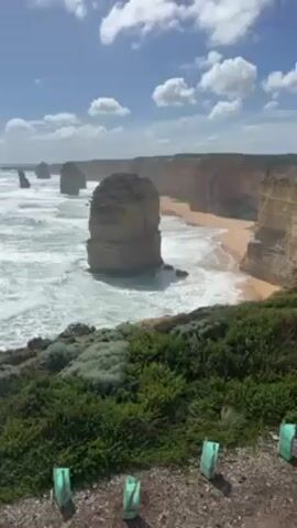 The video shows a scenic view of the Twelve Apostles rock formations on the coast of Australia. The ocean waves crash against the base of the formations and the adjacent sandy beach. The clifftop viewpoint is covered in green vegetation. The sky is blue with scattered clouds. The wind is blowing hard and making noise. Several short, green posts are in the foreground.