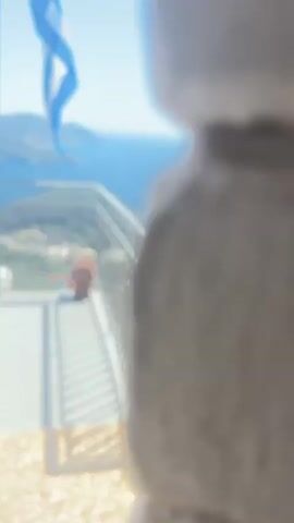 The video shows a panoramic view of a coastal landscape from a platform with a metal railing. A Greek flag waves from a flagpole on the left. A red backpack sits on the platform near the railing. The background features a bay with turquoise water, boats, and lush green hillsides dotted with trees and buildings. The sky is clear and blue. The video appears to be a short, static shot capturing the scenic view.