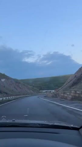 The video is taken from the interior of a vehicle driving on a paved road. The road curves to the right, with hills on either side. The sky is a clear blue with scattered clouds. The sun appears to be low on the horizon, suggesting it is either dawn or dusk. On the right side of the road, there is a stone wall and a house with a red roof is visible in the distance. The overall impression is of a scenic drive through a rural or mountainous area.