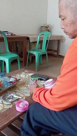 An older man with short gray hair and a light complexion, wearing an orange long-sleeved shirt and dark pants, sits in a chair in front of a wooden table. He is holding a pink thermos with a handle and pouring a clear liquid from it into a glass. On the table, there is an ashtray with cigarette butts and ash, a pink lid, a pack of cigarettes, a lighter, and several glasses. In the background, there are two green plastic chairs and two wooden tables, with a wall behind them. The room appears to be indoors.