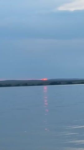 The video shows a wide shot of a body of water. In the distance, the sun is setting behind a silhouetted landscape of hills and trees. The sun's rays are reflecting on the water, creating a bright red and pink streak. The sky is cloudy with a pale blue hue. The water is calm with small ripples. The bottom of the frame shows the rocky edge of the water.