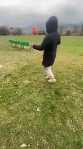 The video shows a person juggling a soccer ball on a grass field. The person is wearing a black hooded jacket, light-colored pants, and tennis shoes. They are standing in a field with scattered debris, including pieces of cardboard and a small can. In the background, there are several green chairs lined up, trees, and a distant mountain range under a cloudy sky. The person is facing away from the camera, using their feet and legs to keep the ball in the air.